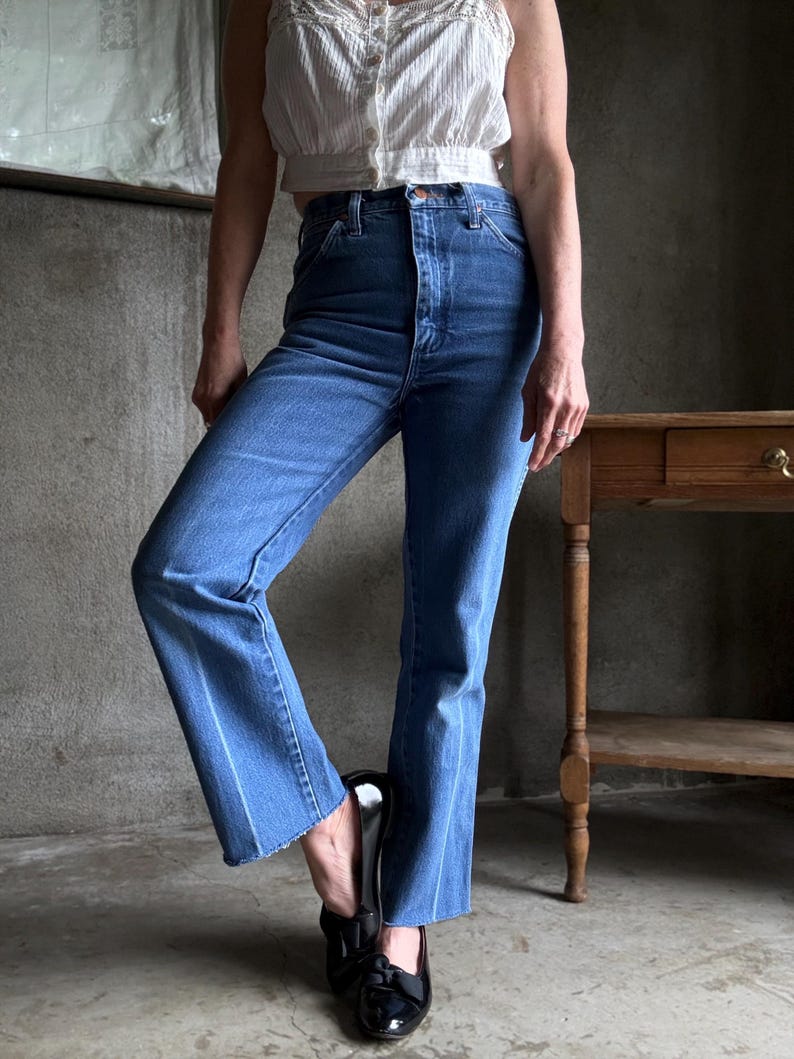 May include: Blue denim jeans with a high waist and flared leg. The jeans have a classic five-pocket design and a light wash. The model is wearing a white top and black shoes. The jeans are a vintage style.