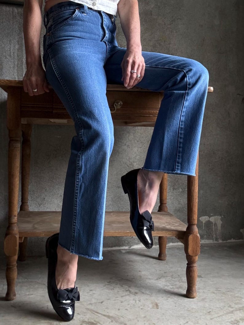 May include: Blue denim jeans with a raw hem, worn with black patent leather flats with a bow. The jeans are high-waisted and the flats have a small heel. The background is a neutral colour.