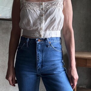 May include: A white, button-down crop top with a square neckline and crochet detailing, paired with high-waisted, blue denim jeans. The top has a textured, vertical stripe pattern. The jeans have a classic five-pocket design.