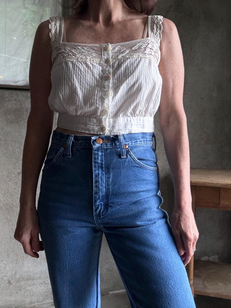 May include: A white, button-down crop top with a square neckline and crochet detailing, paired with high-waisted, blue denim jeans. The top has a textured, vertical stripe pattern. The jeans have a classic five-pocket design.