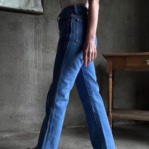 May include: Light blue denim jeans with a flared leg, featuring a classic five-pocket design and a high waist. The jeans have a contrasting white stitch detail along the seams. The person is wearing black loafers.