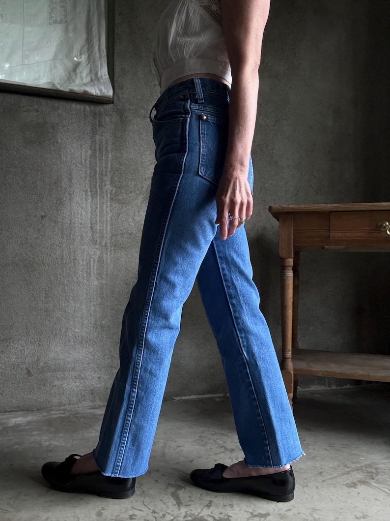 May include: Light blue denim jeans with a flared leg, featuring a classic five-pocket design and a high waist. The jeans have a contrasting white stitch detail along the seams. The person is wearing black loafers.