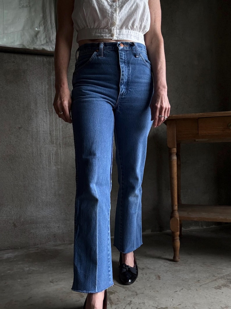 May include: Medium-wash blue denim jeans with a high waist and flared leg. The jeans have a button and zip fastening. The model is wearing a white cropped top and black shoes. The background is a neutral grey.