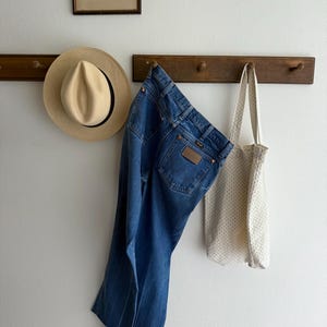 May include: A cream-coloured tote bag with a small polka dot pattern, blue denim jeans, and a light-coloured straw hat hang on a wooden rack. A framed black and white print hangs above the rack.
