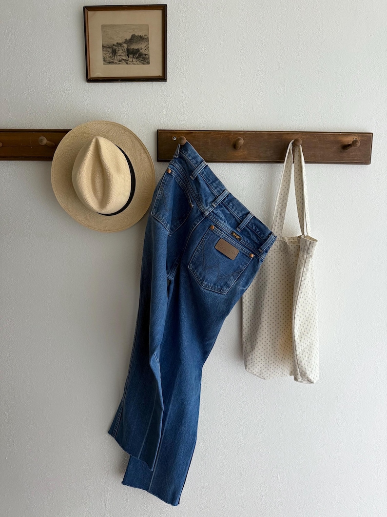 May include: A cream-coloured tote bag with a small polka dot pattern, blue denim jeans, and a light-coloured straw hat hang on a wooden rack. A framed black and white print hangs above the rack.