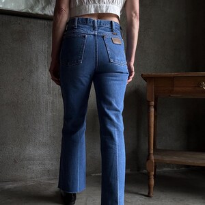 May include: Blue denim jeans with a classic fit and a slight flare at the hem. The jeans feature a brown leather patch on the back pocket and a traditional five-pocket design. The jeans are paired with a white crop top.
