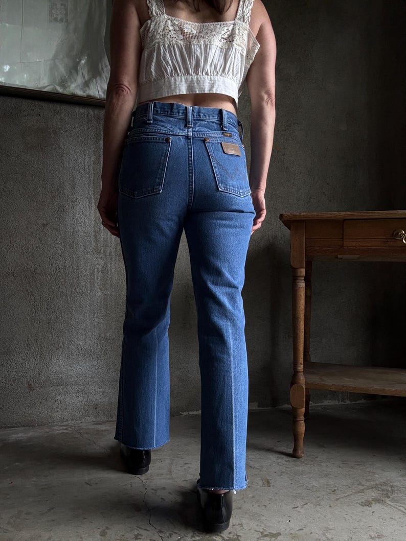 May include: Blue denim jeans with a classic fit and a slight flare at the hem. The jeans feature a brown leather patch on the back pocket and a traditional five-pocket design. The jeans are paired with a white crop top.