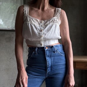 May include: A white, vintage-style tank top with lace detailing and button closure, paired with high-waisted, dark blue denim jeans. The top has a textured, vertical stripe pattern. The jeans have a classic five-pocket design.
