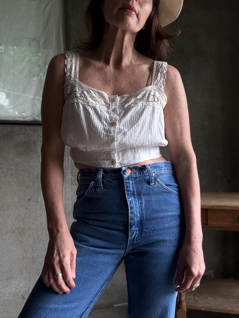 May include: A white, vintage-style tank top with lace detailing and button closure, paired with high-waisted, dark blue denim jeans. The top has a textured, vertical stripe pattern. The jeans have a classic five-pocket design.
