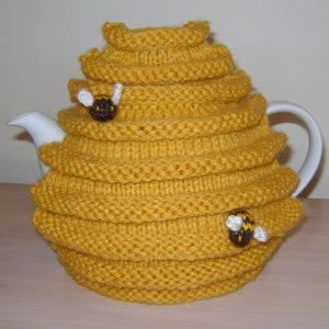 May include: A yellow knitted tea cosy in the shape of a beehive, with a white teapot inside. The cosy has a layered design and is decorated with two small bee appliqués. The cosy is designed to keep tea warm.
