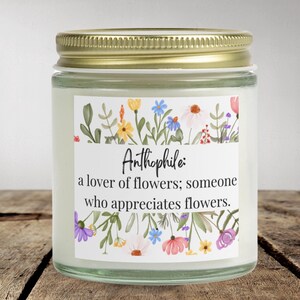 May include: A white candle jar with a gold lid and a floral patterned label. The label reads "Anthophile: a lover of flowers; someone who appreciates flowers."
