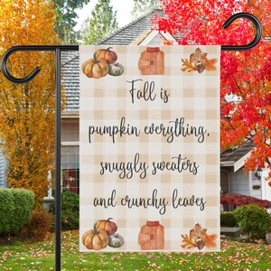 Könnte beinhalten: Gartenflagge zum Thema Herbst mit kariertem Hintergrund. Die Flagge zeigt eine Aquarell-Illustration von Kürbissen, einem Pullover und Blättern. Der Text auf der Flagge lautet "Fall is pumpkin everything, snuggly sweaters and crunchy leaves."