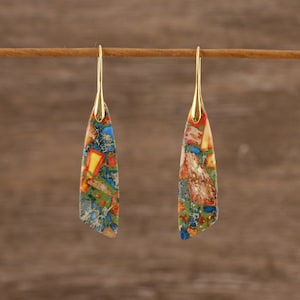 Puede incluir: Un par de pendientes colgantes con ganchos dorados. Los pendientes tienen un colorido patrón de mosaico en tonos de azul, verde, amarillo y rojo. Los pendientes cuelgan sobre un fondo marrón.