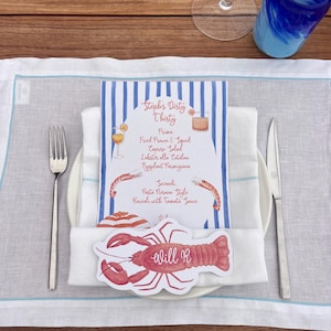 Peut inclure: Mise en place avec un menu pour "Steph's Dirty Thirty" et un marque-place en forme de homard. Le menu présente un design à rayures bleues et blanches et répertorie des plats comme les crevettes et calmars frits. Une fourchette, un couteau et un verre sont également visibles.