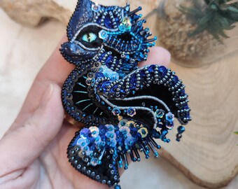 Puede incluir: Un broche de cuentas azules y negras con un diseño detallado de una criatura mítica con un ojo grande. El broche está hecho con lentejuelas y cuentas y tiene un efecto tridimensional.