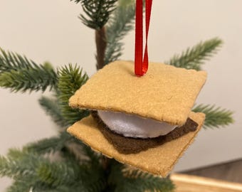 May include: A handmade felt s'mores ornament with a red ribbon for hanging. The ornament features two tan graham cracker squares, a dark brown chocolate layer, and a white marshmallow center. The ornament is displayed against a backdrop of a small artificial Christmas tree.