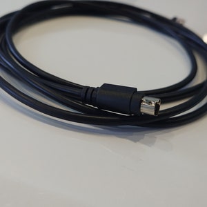 May include: A black S-Video cable coiled on a white surface. The cable has a black connector with a silver-coloured metal end. The cable is used to transmit video signals.
