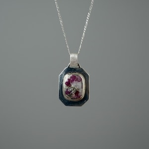 May include: A silver pendant necklace with a rectangular, octagonal frame. The pendant features a central stone with a mix of pink and white hues, and a silver chain. The necklace is displayed against a gray background.
