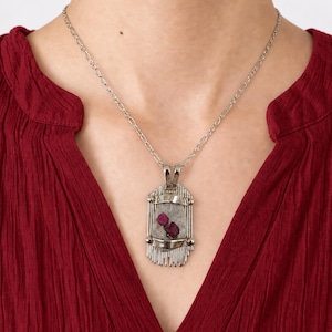 May include: A silver-toned pendant necklace featuring a rectangular metal frame with vertical grooved detailing at the top and bottom. The center holds a translucent, light-colored stone embedded with two small, deep pink, raw-cut ruby crystals.