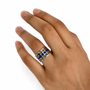 May include: A wide silver band ring featuring a grid-like openwork design. The ring is accented with small, square-shaped inlaid stones in alternating colors of deep blue, black, and green, set within the metallic lattice structure.