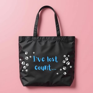 May include: Black tote bag featuring white paw prints and the blue text "I've lost count...". The bag has two black handles and is set against a pink background. A practical accessory for carrying items, perfect for pet owners.