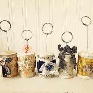 May include: Five decorative card holders with circular wire tops. The holders are made from repurposed spools and adorned with lace, ribbons, and charms. Each holder has a unique design, including a key charm and a blue jewel.