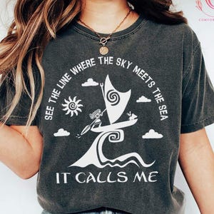 以下が含まれることがあります： ダークヘザーのComfort Colors Tシャツ。白いグラフィックは、太陽、雲、そして「SEE THE LINE WHERE THE SKY MEETS THE SEA IT CALLS ME」のテキストが入った、様式化されたヨットが特徴です。