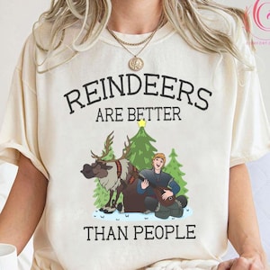 May include: Cream-colored t-shirt with the text "REINDEERS ARE BETTER THAN PEOPLE." The graphic features a cartoon reindeer, a man playing a lute, and Christmas trees. The shirt is labeled "Natural" at the bottom.