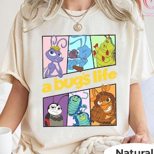 May include: A cream-colored t-shirt featuring a colorful graphic from the animated film "A Bug's Life." The design includes various insect characters in rectangular panels, with the film's title in yellow. The shirt is labeled "Natural" at the bottom.