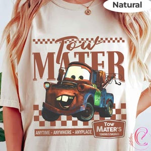 Puede incluir: Una camiseta color crema con una imagen de dibujos animados de Mate de la pel&iacute;cula Cars. El dise&ntilde;o incluye el texto "Tow Mater" en marr&oacute;n, junto con las frases "ANYTIME - ANYWHERE - ANYPLACE" y "TOWING & SALVAGE".
