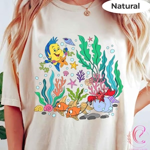 K&ouml;nnte beinhalten: Cremefarbenes T-Shirt mit einer farbenfrohen Unterwasserszene mit Zeichentrickfiguren. Das Design umfasst Fische, eine Krabbe, Seetang und andere Meeresbewohner. Das Wort "Natural" ist oben rechts aufgedruckt.