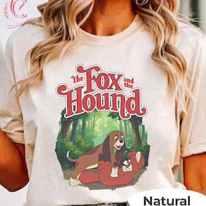 Puede incluir: Camiseta de color natural con un gráfico de "Tod y Toby". El diseño muestra un perro marrón y un zorro rojo en un bosque, con el título en escritura roja arriba. La palabra "Natural" está impresa en la parte inferior.