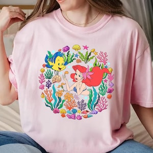 Pu&ograve; includere: T-shirt rosa chiaro con una grafica colorata di Ariel, Flounder ed elementi marini. Il design &egrave; una disposizione circolare di coralli, conchiglie e alghe. Il testo "LIGHT PINK" &egrave; in basso sulla maglietta.