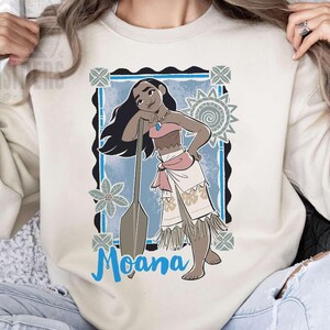 May include: Sand-coloured sweatshirt featuring a graphic of Moana, with a blue background and the word "Moana" in blue script. The design includes a cartoon illustration of Moana with a paddle. The sweatshirt is a neutral colour.