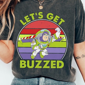 May include: A dark gray t-shirt with a retro-style graphic of Buzz Lightyear from Toy Story. The graphic features a colorful sunset with the text "Let's Get Buzzed".