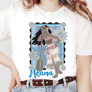 May include: White t-shirt with a graphic of Moana, framed in blue and black. The illustration depicts Moana with long black hair, a pink top, and a white skirt, leaning on a paddle. The word "Moana" is written in blue.