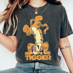 Könnte beinhalten: Ein dunkelgraues T-Shirt mit einem Cartoon-Bild von Tigger aus Winnie Puuh. Tigger steht auf dem Kopf mit seinen Pfoten in der Luft. Der Text "TIGGER" ist in Orange unter dem Bild gedruckt.