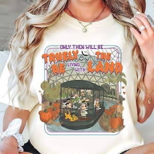 May include: A cream-colored t-shirt featuring a graphic print of cartoon characters on a boat ride through a greenhouse. The text reads ONLY THEN WILL BE TRULY THE LIVING WITH LAND. The design includes orange pumpkins, green foliage, and a spiderweb background.