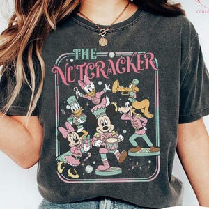 May include: Dark heather t-shirt featuring a colorful graphic of Disney characters in Nutcracker costumes. The design includes Mickey Mouse, Minnie Mouse, Donald Duck, Daisy Duck, and Goofy. The text "THE NUTCRACKER" is displayed above the characters.