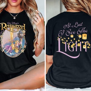 Op de afbeelding: Een zwart t-shirt met een afbeelding op de voorkant van Rapunzel met lang blond haar in een toren, omringd door een bloemenrand en de tekst Walt Disney's Rapunzel. De achterkant toont de tekst At Last I See The Light met zwevende lantaarns.