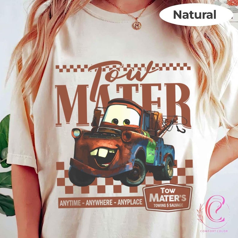 Puede incluir: Una camiseta color crema con una imagen de dibujos animados de Mate de la pel&iacute;cula Cars. El dise&ntilde;o incluye el texto "Tow Mater" en marr&oacute;n, junto con las frases "ANYTIME - ANYWHERE - ANYPLACE" y "TOWING & SALVAGE".
