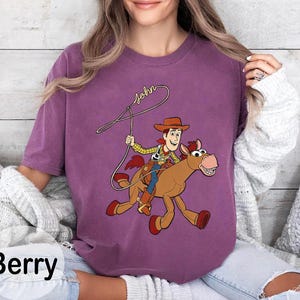 May include: Berry-colored t-shirt featuring a cartoon cowboy riding a horse, with the name "John" written above. The cowboy wears a hat, vest, and blue jeans. The horse is brown with red accents.