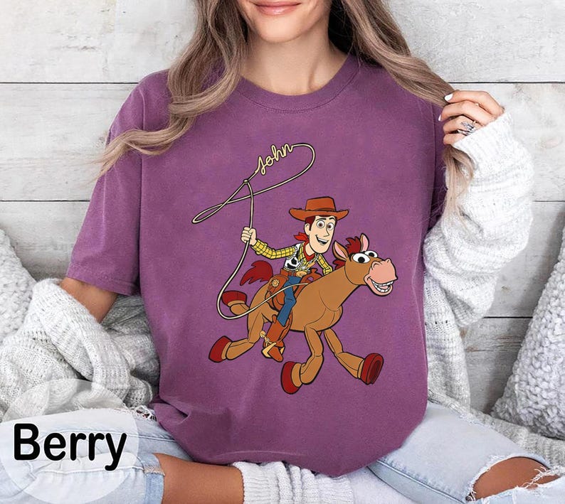 May include: Berry-colored t-shirt featuring a cartoon cowboy riding a horse, with the name "John" written above. The cowboy wears a hat, vest, and blue jeans. The horse is brown with red accents.