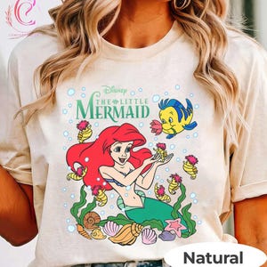 Puede incluir: Camiseta beige con un gr&aacute;fico colorido de Ariel de La Sirenita de Disney. El dise&ntilde;o incluye a Ariel con cabello rojo, Flounder y caballitos de mar, rodeados de burbujas y conchas marinas. Las palabras "Disney The Little Mermaid" est&aacute;n arriba.