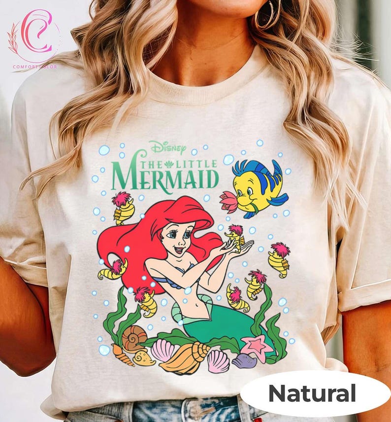 Puede incluir: Camiseta beige con un gr&aacute;fico colorido de Ariel de La Sirenita de Disney. El dise&ntilde;o incluye a Ariel con cabello rojo, Flounder y caballitos de mar, rodeados de burbujas y conchas marinas. Las palabras "Disney The Little Mermaid" est&aacute;n arriba.