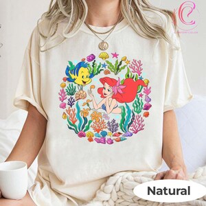 Pu&ograve; includere: T-shirt color crema con una grafica colorata di Ariel de La Sirenetta con Flounder, circondata da coralli, conchiglie e alghe. La parola "Natural" &egrave; stampata in basso.