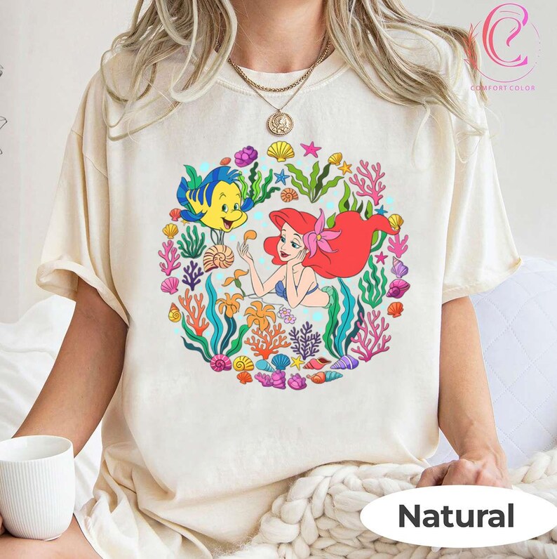 Pu&ograve; includere: T-shirt color crema con una grafica colorata di Ariel de La Sirenetta con Flounder, circondata da coralli, conchiglie e alghe. La parola "Natural" &egrave; stampata in basso.