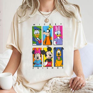 May include: A cream-colored t-shirt with a graphic print of Mickey Mouse and his friends. The graphic features Mickey Mouse, Minnie Mouse, Donald Duck, Goofy, Pluto, and Daisy Duck in colorful squares. The text "Mickey and Friends" is printed below the graphic.