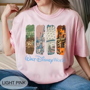 May include: Light pink t-shirt with a Walt Disney World graphic. The design features four vertical rectangular images with illustrations of the Tree of Life, a castle, a geometric structure, and the Tower of Terror. The words "Walt Disney World" are written in light blue.