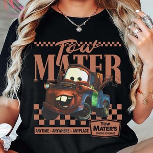 Puede incluir: Camiseta negra con una imagen de dibujos animados de Mate de la pel&iacute;cula Cars. El dise&ntilde;o incluye el texto "Tow Mater" encima del personaje y "ANYTIME - ANYWHERE - ANYPLACE" debajo. Un peque&ntilde;o letrero dice "Tow Mater's Towing & Salvage."